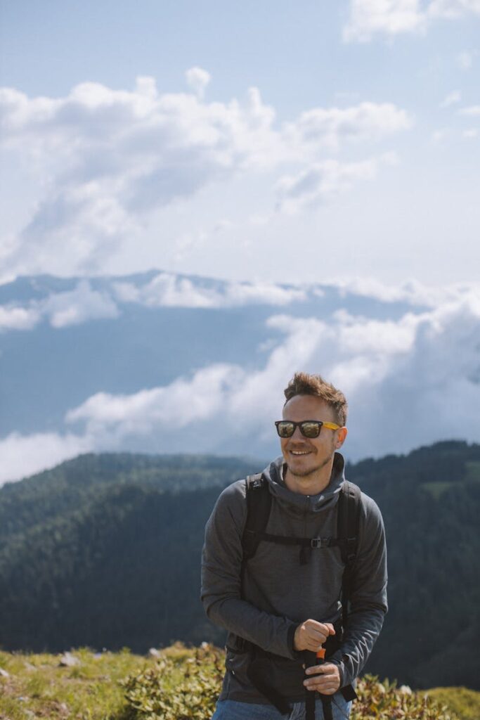 A man with sunglasses smiles while hiking in the mountains under a cloudy sky.